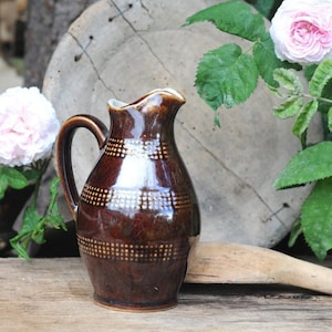 May include: A brown ceramic pitcher with a handle and a decorative dotted pattern. The pitcher has a glossy finish and a white rim. Pink roses and green leaves are in the background.
