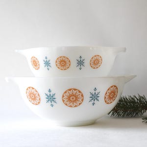 Pode incluir: Duas tigelas brancas empilhadas, com um design de floco de neve e floral em laranja e azul-petróleo. As tigelas têm uma forma clássica com uma alça em cada lado, perfeitas para uso na cozinha.