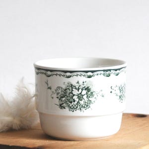May include: White ceramic cup with a green floral design. The cup has a stacked design and a dark green trim. The floral pattern includes detailed leaves and flowers. The cup is on a wooden surface.