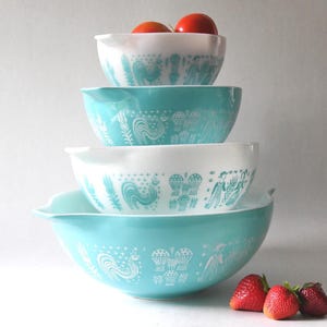 May include: A set of four turquoise and white mixing bowls with a rooster and floral design. The bowls are stacked on top of each other, with the largest bowl at the bottom and the smallest bowl at the top. There are two red apples on top of the smallest bowl.