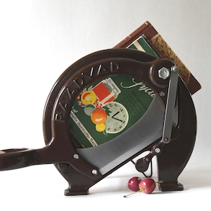 Puede incluir: Una rebanadora de alimentos vintage de metal marrón con una cuchilla curva y un mango. La rebanadora tiene la palabra "BAZAD". Es visible una etiqueta verde y blanca con ilustraciones de frutas. Dos cerezas descansan cerca de la cuchilla.