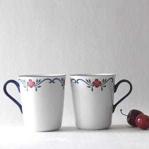 May include: Two white ceramic mugs with blue and green floral designs and blue handles. The mugs are sitting on a white surface.