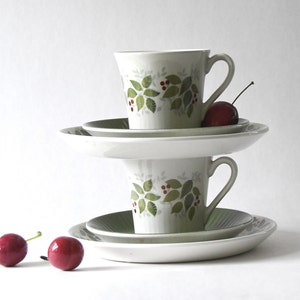 May include: Two stacked teacups and saucers with a leaf and berry design. The cups are white with green leaves and red berries. Each cup sits on a matching saucer. Two red cherries are in the foreground.