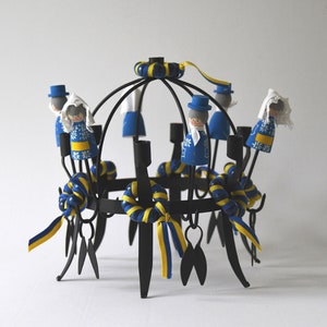 Puede incluir: Un candelabro de metal negro con un dise&ntilde;o circular, adornado con peque&ntilde;as figuras de madera vestidas con trajes azules y blancos. Cintas y detalles amarillos y azules a&ntilde;aden color. Las figuras est&aacute;n dispuestas alrededor de los candelabros.