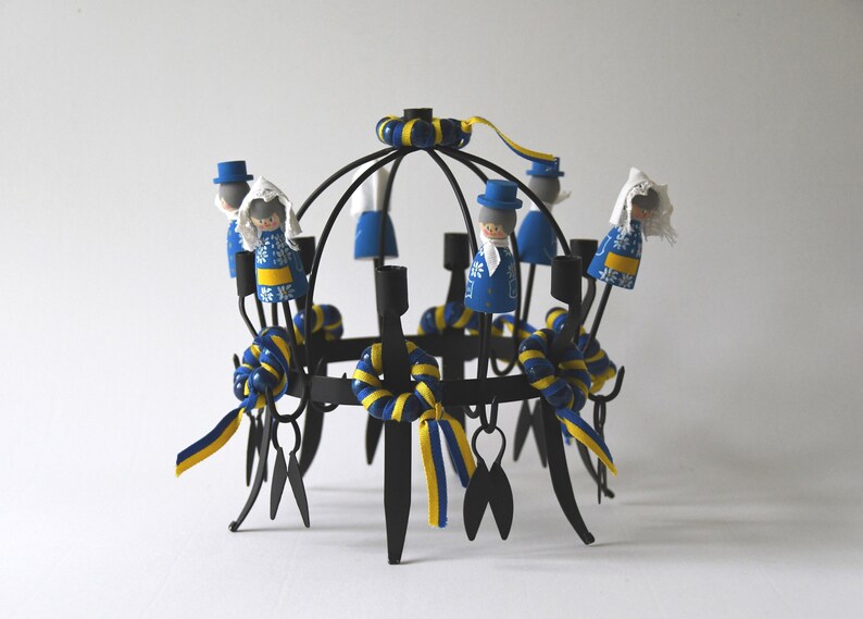Puede incluir: Un candelabro de metal negro con un dise&ntilde;o circular, adornado con peque&ntilde;as figuras de madera vestidas con trajes azules y blancos. Cintas y detalles amarillos y azules a&ntilde;aden color. Las figuras est&aacute;n dispuestas alrededor de los candelabros.