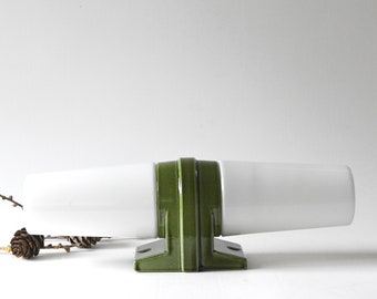 Puede incluir: Un objeto de cerámica horizontal con dos formas cilíndricas blancas y cónicas conectadas por una base rectangular de color verde oliva con un arco central elevado. La base incluye dos orificios circulares visibles en sus pies planos.