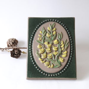 May include: A ceramic tile featuring a raised design of yellow flowers within an oval frame. The tile is bordered by a green frame with a beaded edge.
