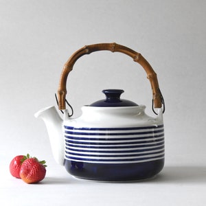 Könnte beinhalten: Weiß-blaue Keramik-Teekanne mit horizontalen Streifen, Bambusgriff und marineblauem Deckel. Zwei rote Erdbeeren liegen links. Ein stilvolles Küchenaccessoire.