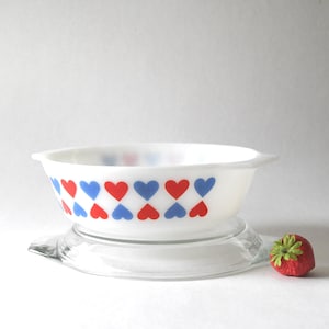 Op de afbeelding: Witte Pyrex-schaal met een patroon van rode en blauwe harten. De schaal staat op een heldere glazen standaard. Een rode aardbei met groene bladeren ligt naast de schaal.