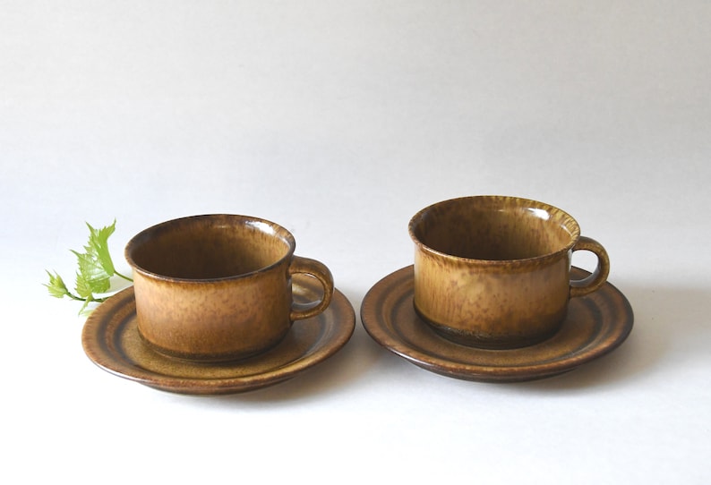 May include: Two brown ceramic teacups and saucers. The cups have a textured, speckled glaze. The saucers have a brown and white striped pattern.