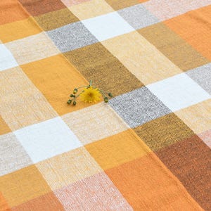 May include: A woven fabric featuring a large-scale checkered pattern in shades of orange, yellow, white, and grey. The textile has a visible textured weave and is decorated with a single small yellow flower placed near the center of the pattern.