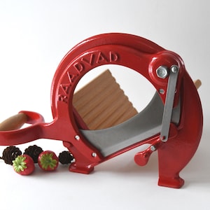 Puede incluir: Un cortador de alimentos de hierro fundido rojo con un mango de madera y una cuchilla plateada. El cortador está etiquetado como "RAADVAAD" y tiene una guía de madera para cortar.
