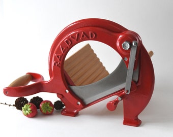 May include: A red cast iron food slicer with a wooden handle and a silver blade. The slicer is labelled "RAADVAAD" and has a wooden guide for slicing.