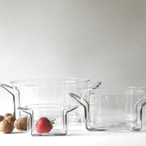 Può includere: Tre ciotole di vetro trasparente con manici in metallo, annidate l'una nell'altra. La ciotola più piccola contiene una singola fragola rossa. Le ciotole sono su una superficie bianca.