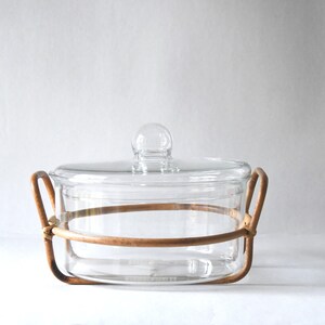 May include: A clear glass casserole dish with a matching glass lid featuring a round knob handle. The dish is held within a light brown, circular rattan or wicker frame that includes two integrated side handles for lifting and carrying.