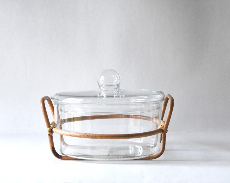 May include: A clear glass casserole dish with a matching glass lid featuring a round knob handle. The dish is held within a light brown, circular rattan or wicker frame that includes two integrated side handles for lifting and carrying.