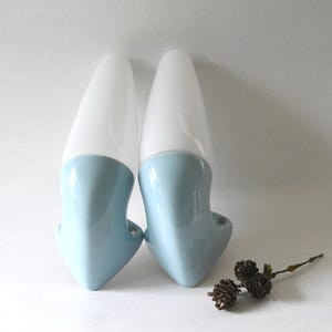 May include: A pair of vintage wall sconces. Each sconce has a white, elongated, cone-shaped shade and a light blue, curved ceramic base. The sconces are mounted on a white wall, offering a minimalist aesthetic.