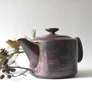 Pode incluir: Uma chaleira de cerâmica com esmalte bordô escuro, com tampa e alça. Um raminho de pinheiro e uma pinha acompanham a chaleira. É um artigo de cozinha.