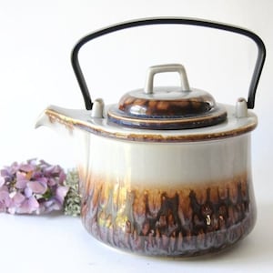 Può includere: Una teiera in ceramica con una sezione superiore bianco sporco lucida che sfuma in un motivo a smalto gocciolante marrone scuro alla base. Include un coperchio abbinato con una piccola maniglia e un manico ad arco in metallo nero sui lati.