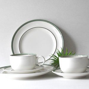 May include: White ceramic dinnerware set with a green and black trim. The set includes two teacups, saucers, and plates. A large dinner plate is in the background. The set is arranged on a white surface.