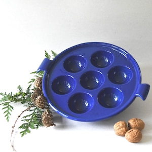 May include: A round, glossy cobalt blue ceramic escargot dish featuring seven circular indentations and two side handles. The dish is accompanied by a small sprig of green cedar with tiny brown cones and three whole walnuts placed nearby.