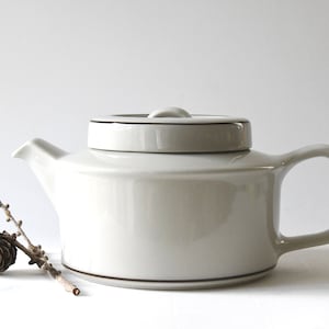 Op de afbeelding: Een witte theepot van keramiek met een bruine rand rond de deksel en de basis. De theepot heeft een rond lichaam en een gebogen handvat.