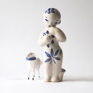 May include: A white ceramic figurine of a girl with blue floral designs, holding a bouquet of flowers. A small white fluffy lamb with a blue collar stands beside her.
