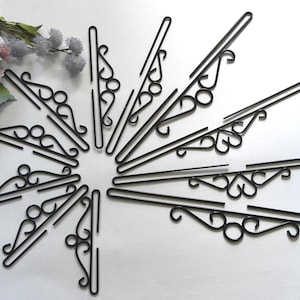 Könnte beinhalten: Schwarze dekorative Wandhalterungen aus Metall, kreisförmig angeordnet. Jede Halterung hat einen geraden Arm mit dekorativen Schnörkeln und einen gebogenen Haken. Die Halterungen werden auf einer weißen Oberfläche mit floralen Akzenten im Hintergrund präsentiert.