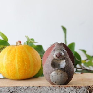 May include: A small, dark brown ceramic figurine shaped like a stylized animal with a rounded body, a lighter speckled belly, and a simple carved face. The figure has a smooth, matte texture and sits upright, featuring a small nose and a subtle smile.