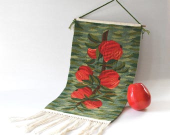 May include: A woven wall hanging with a botanical design featuring red flowers and green leaves against a green background. The tapestry has a fringe at the bottom and is suspended by a wooden rod and green cord. A red ceramic decorative object is next to the tapestry.