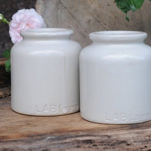May include: Two off-white ceramic jars with a smooth, cylindrical body and a short, rounded rim. Each jar features the embossed text LAB Lagny near the base. The vessels have a simple, functional design with a glossy finish and a wide opening.