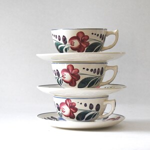 May include: Stacked vintage teacups and saucers. Each cup and saucer is white with a floral design featuring red flowers, green leaves, and blue accents. The cups have a curved handle and are stacked on top of each other.