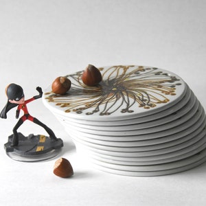 Può includere: Una pila di sottobicchieri in ceramica bianca con un motivo floreale oro e marrone, accanto a una statuina di una supereroina in tuta rossa e nera. Tre nocciole sono sparse intorno ai sottobicchieri.