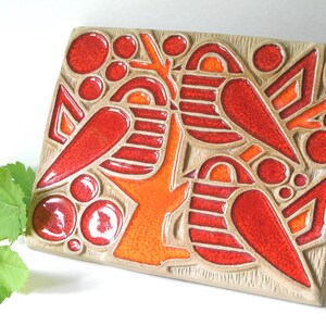 May include: Rectangular ceramic tile with a beige background and a vibrant design in red and orange. The design features abstract shapes, including circles, triangles, and bird-like forms, creating a striking visual pattern. The tile has a glossy finish.