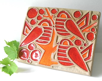 May include: Rectangular ceramic tile with a beige background and a vibrant design in red and orange. The design features abstract shapes, including circles, triangles, and bird-like forms, creating a striking visual pattern. The tile has a glossy finish.