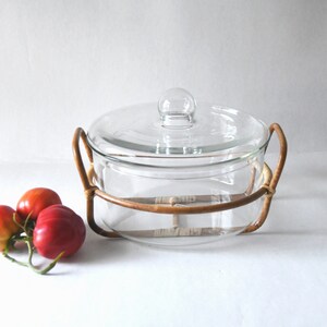 May include: A clear glass casserole dish with a matching lid and a round glass knob handle, set inside a brown rattan frame with two side handles. Three red tomatoes with green stems are positioned to the left of the glass dish.