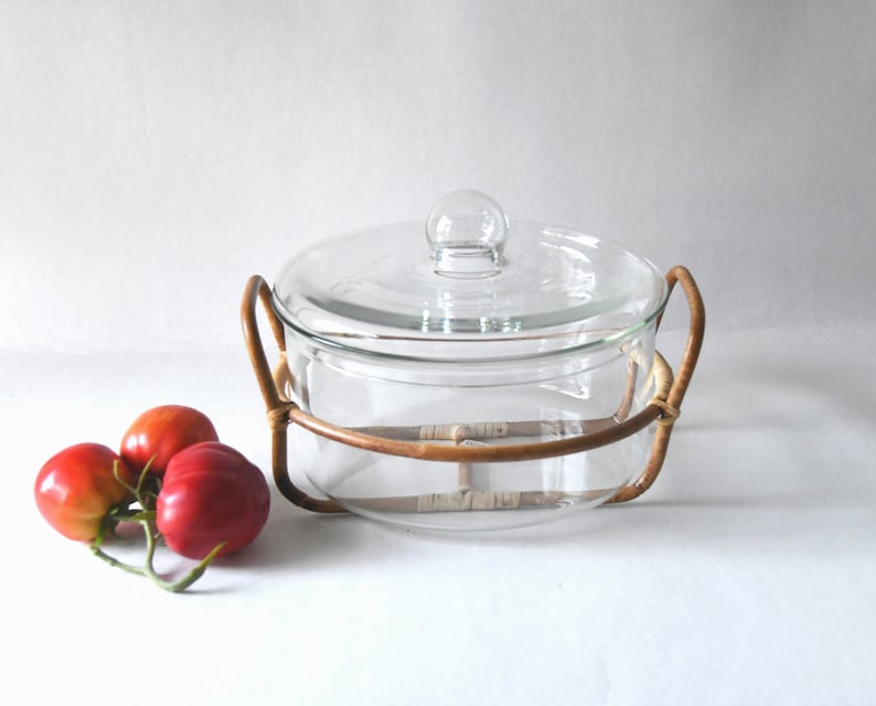 May include: A clear glass casserole dish with a matching lid and a round glass knob handle, set inside a brown rattan frame with two side handles. Three red tomatoes with green stems are positioned to the left of the glass dish.