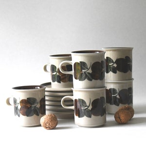 May include: A set of six brown and white ceramic coffee mugs with a leafy pattern. The mugs are stacked in two piles of three. There are two walnuts on the table next to the mugs.