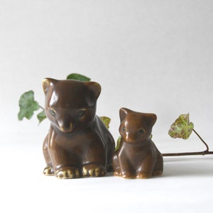 May include: Two glossy brown ceramic bear figurines of different sizes, positioned side by side. The larger bear is on the left and the smaller bear is on the right. Both figurines feature smooth, reflective surfaces and pointed ears.