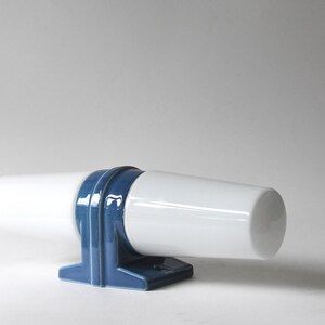 Op de afbeelding: Een witte keramische wandlamp met een cilindrische kap en een blauwe decoratieve steun. De lamp heeft een geometrisch ontwerp en is gemonteerd op een rechthoekige basis. De armatuur is ontworpen voor gebruik binnenshuis.