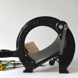 Puede incluir: Cortador de pan de hierro fundido negro de la marca RADWAY con base de madera y cuchilla plateada.