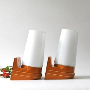 Peut inclure: Deux lampes de table assorties avec des abat-jours cylindriques blancs et des bases orange-brun. Les bases ont un design en gradins avec des rainures horizontales. Une branche de baies rouges et de feuilles vertes est placée près d'une lampe.
