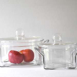 Könnte beinhalten: Zwei transparente Glas-Auflaufformen mit Deckeln und Metallgriffen. Die größere Form enthält zwei rote Tomaten. Beide Formen stehen auf Metallständern.