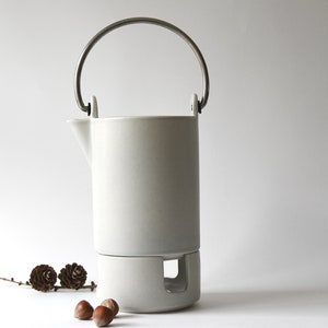 Può includere: Teiera in ceramica bianca con manico argentato e scaldavivande integrato. La teiera è appoggiata su una superficie bianca con alcuni piccoli pigne marroni e nocciole.