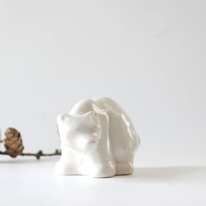 May include: A small, white ceramic polar bear figurine. The bear is in a seated position with its head slightly bowed. Two pine cones and a twig are in the background. The figurine is a decorative item.