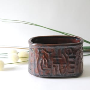 Puede incluir: Un recipiente rectangular de cerámica con un exterior marrón rojizo oscuro y un interior oscuro. El recipiente tiene una superficie texturizada con un diseño en relieve. El fondo incluye elementos decorativos blancos y verdes.
