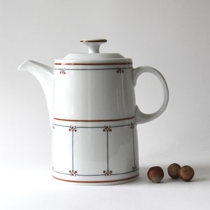 Puede incluir: Una cafetera de cerámica blanca con tapa y asa. La cafetera está decorada con líneas marrones y azules y pequeños puntos. Tres avellanas están al lado de la cafetera.