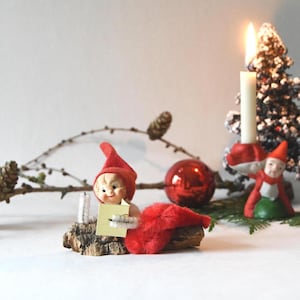May include: A small elf figurine with a red hat and fuzzy red body sits on a piece of bark, holding a small yellow book. Beside it, a second elf figurine stands on a green base, holding a white candle with a lit flame, near two snowy trees.