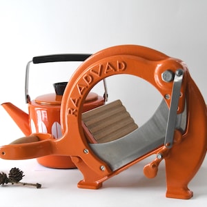 Puede incluir: Cortadora de alimentos vintage naranja con una cuchilla plateada y una tabla de cortar de madera. La cortadora está etiquetada como "RÅDVAD".