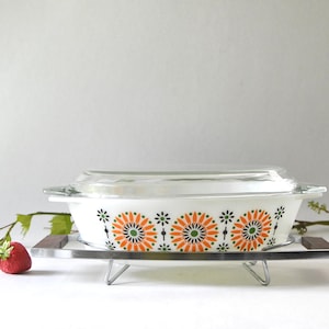 Könnte beinhalten: Weißer Auflaufform mit transparentem Glasdeckel, verziert mit einem orangefarbenen, grünen und schwarzen Blumenmuster. Die Form steht auf einem Metallständer mit Holzakzenten. Vintage Küchenutensil.